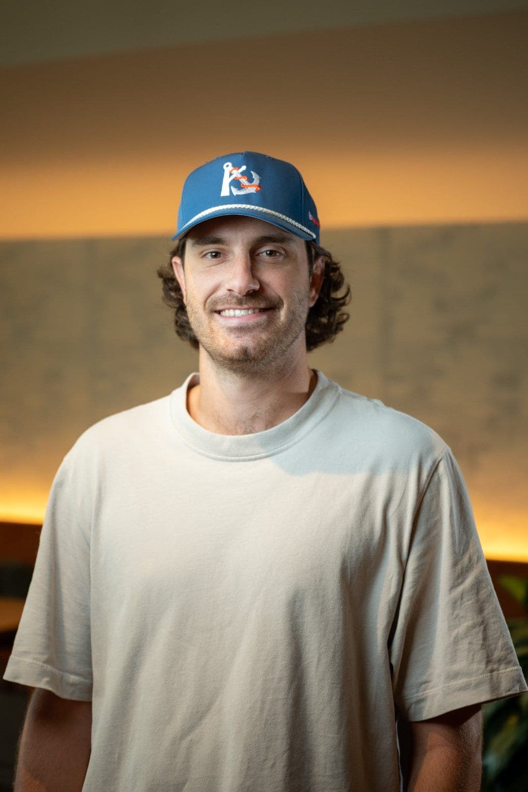 Professional headshot of a smiling man wearing a cap and light shirt in an indoor workplace setting.