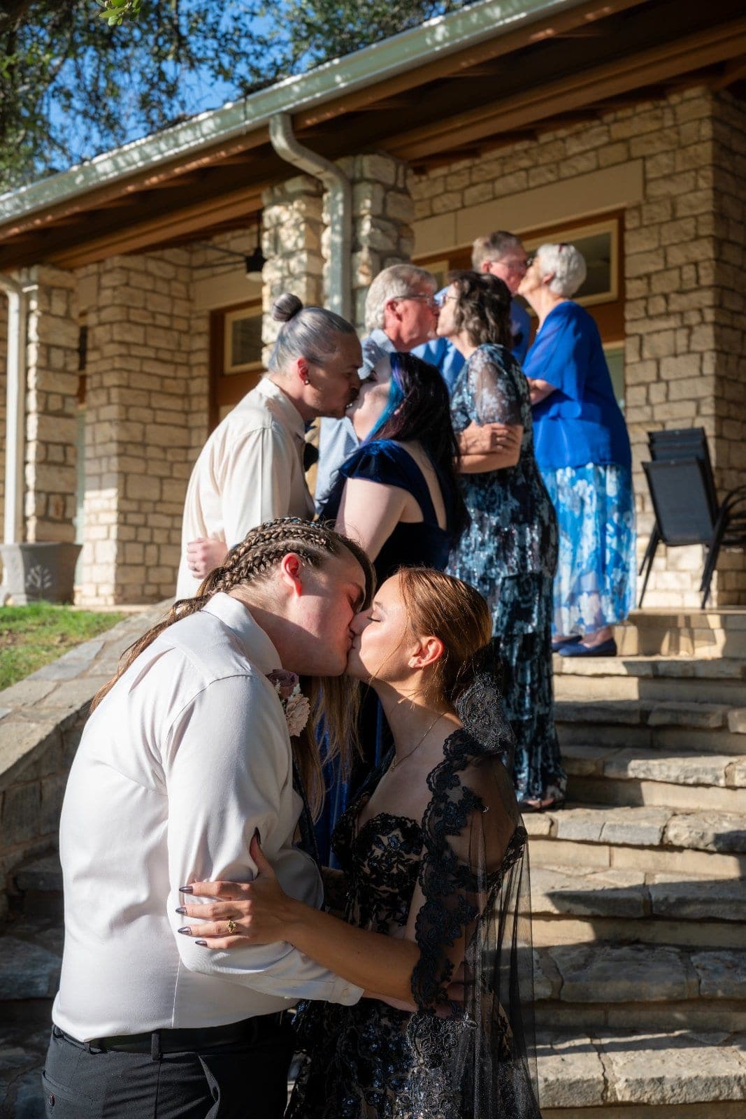 Newlywed couple kissing in the foreground while family members celebrate together behind them at an outdoor Marble Falls wedding.