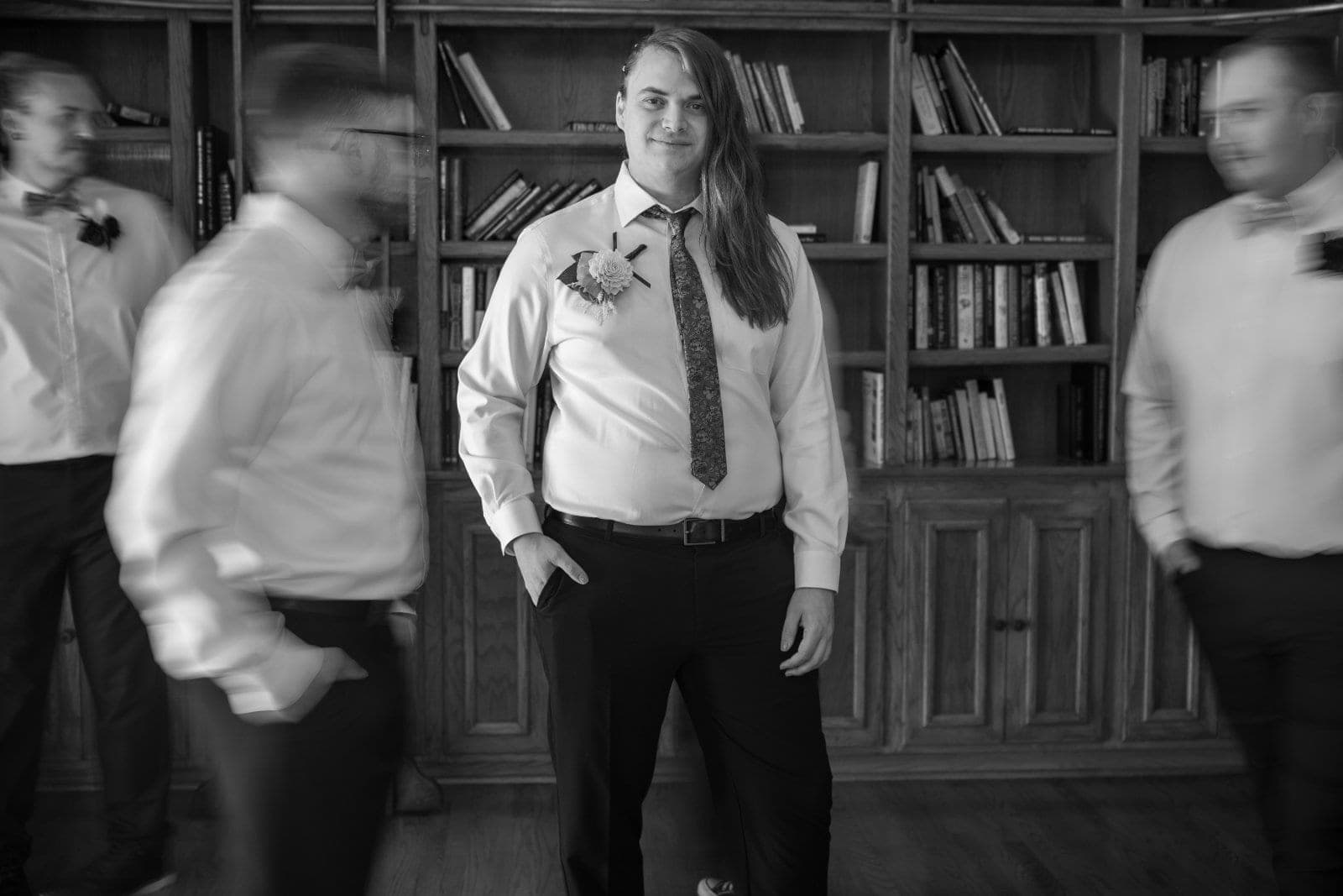 Black-and-white creative wedding portrait of the groom standing in focus while other members of the wedding party move around him.