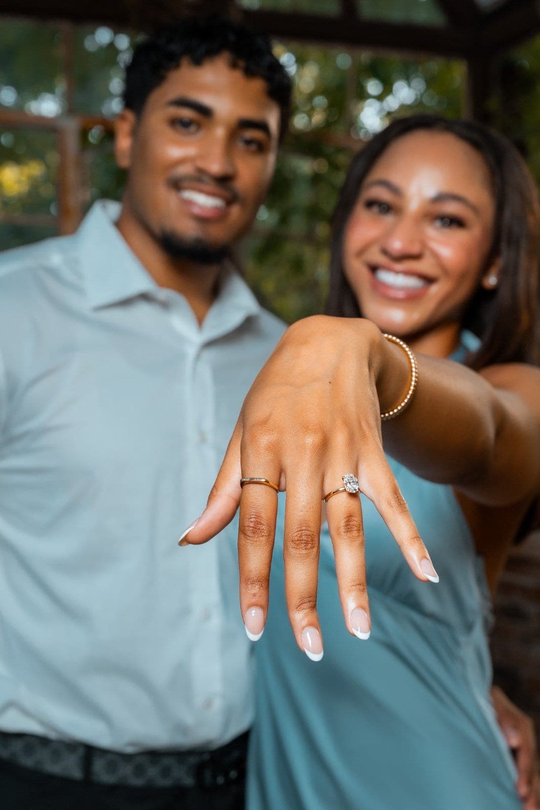 Close-up of a newly engaged woman showing her engagement ring with her partner smiling beside her after a surprise proposal.