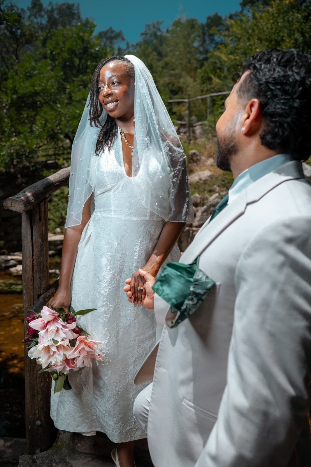 Bride in a veil holding her bouquet and the groom’s hand beside a rustic wooden railing during their outdoor wedding portraits.