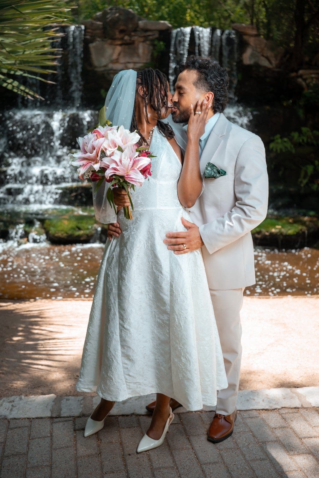 Newlywed couple sharing a kiss while the bride holds a floral bouquet in front of a waterfall in Austin, Texas.