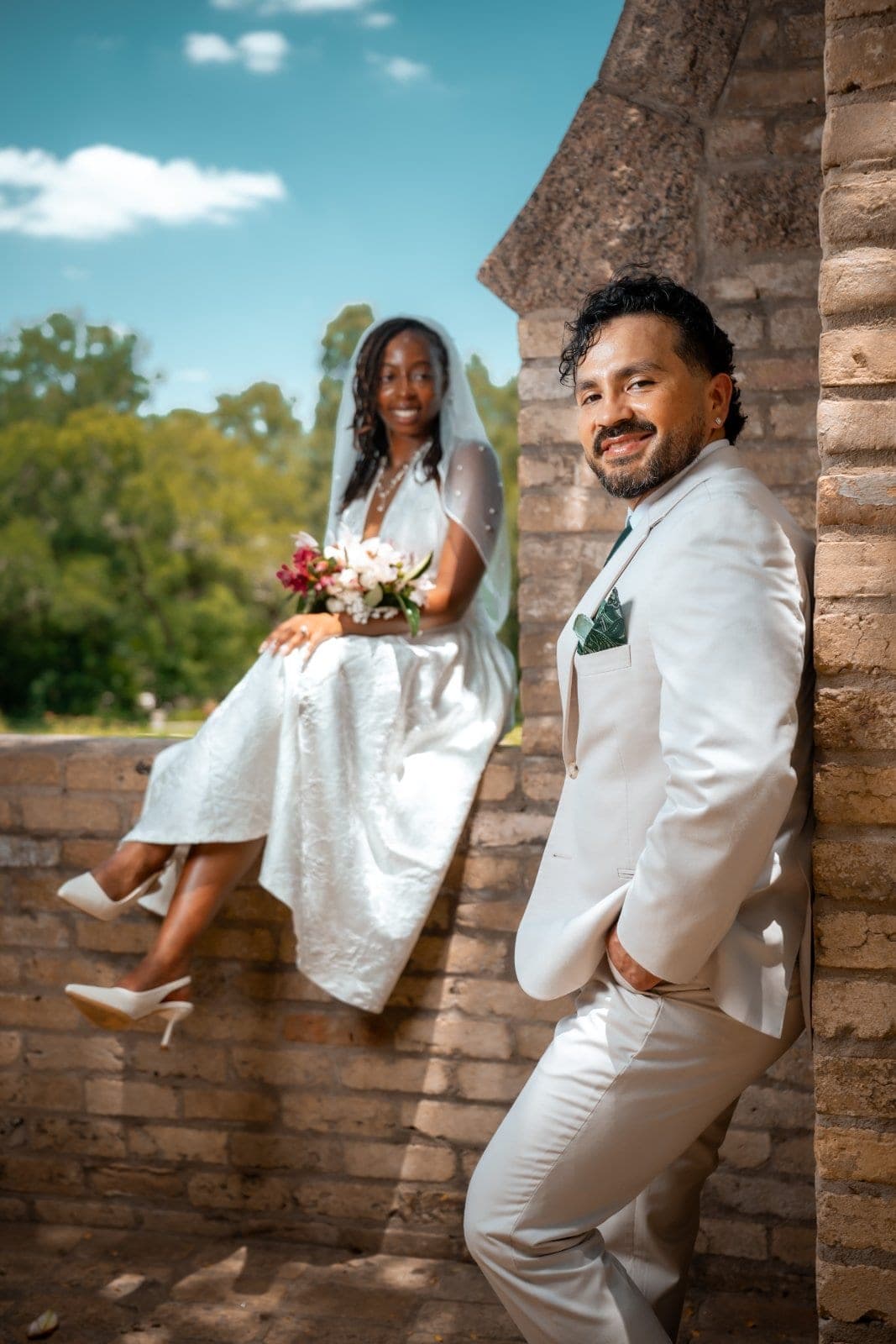 Groom posing beside a stone arch with the bride seated in the background holding her wedding bouquet.