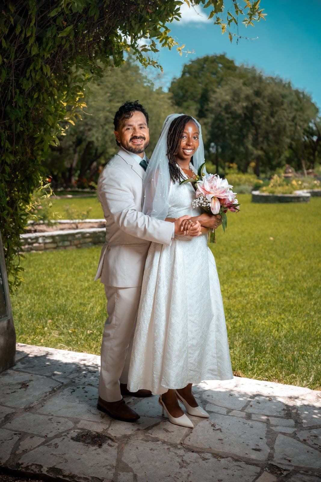 Bride and groom posing together with a floral bouquet in a sunlit garden surrounded by greenery in Texas.