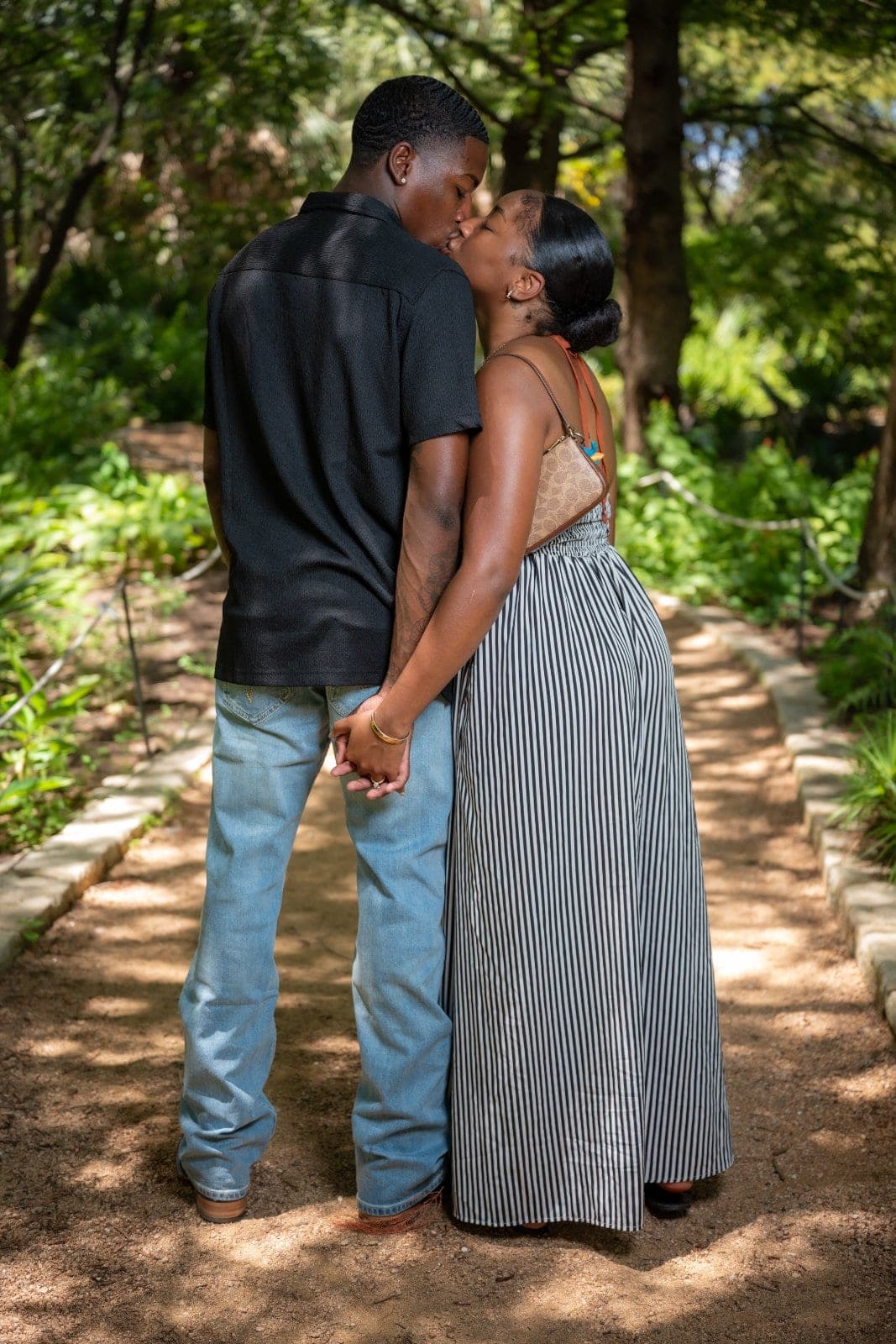 Newly engaged couple holding hands and sharing a kiss while walking along a lush garden path in Austin.