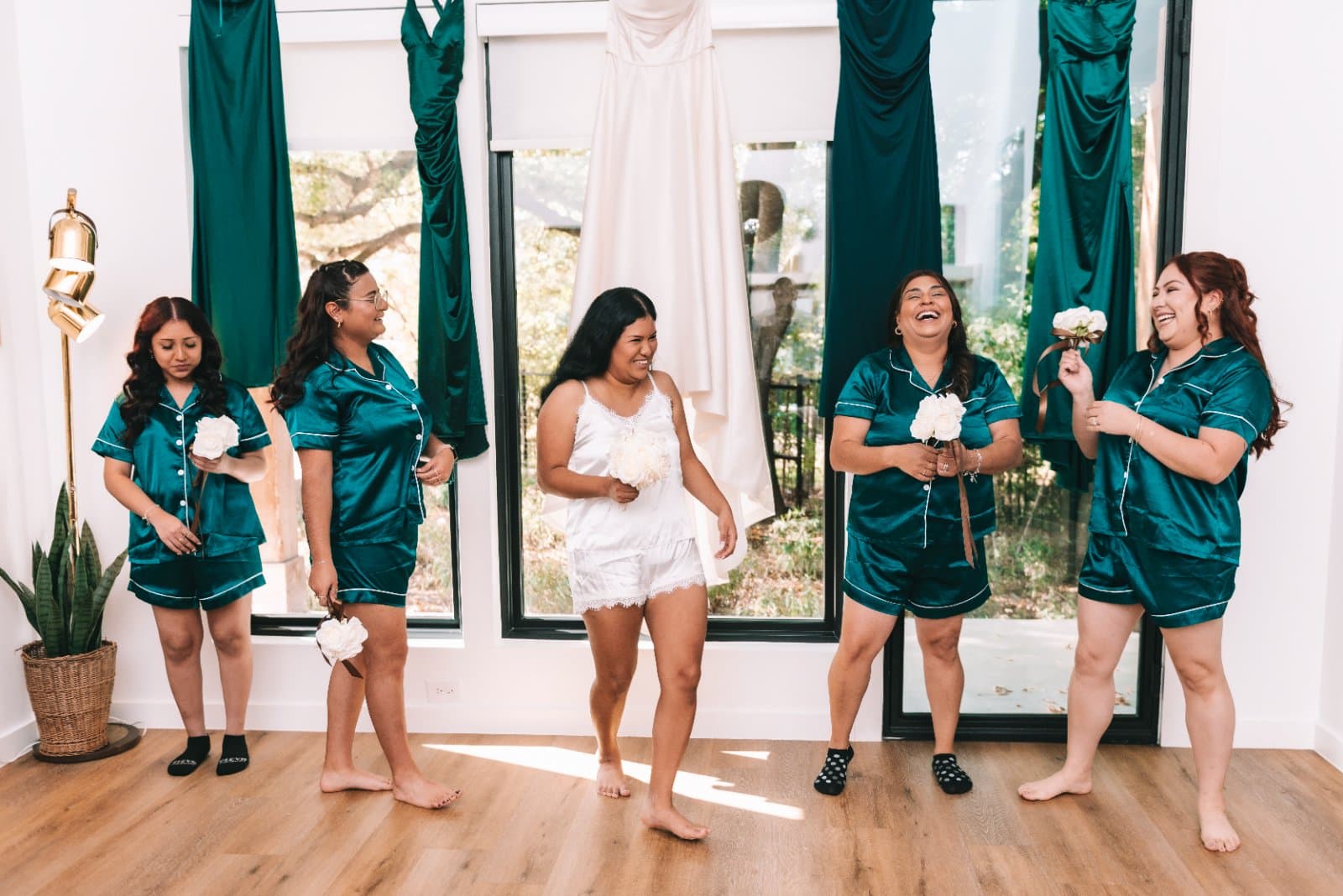 Bridesmaids in matching satin pajamas holding white bouquets beside the bride’s wedding dress in Austin, Texas.